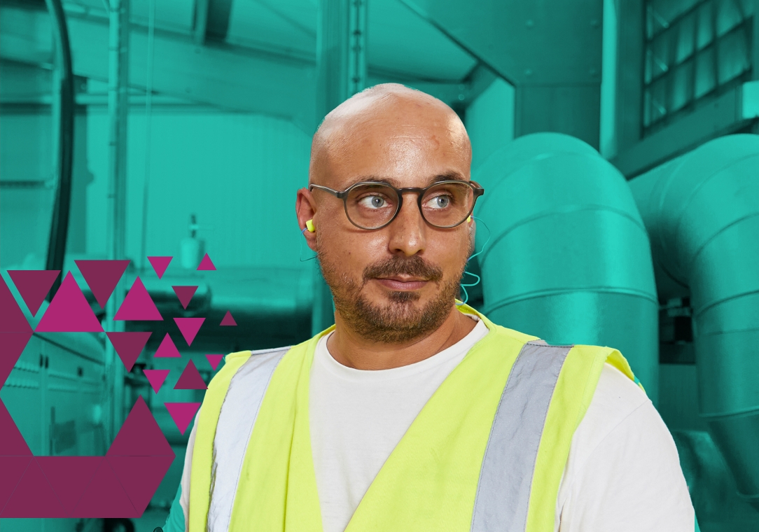 A bald man with a beard wearing glasses and a yellow safety vest stands in an industrial setting with green machinery in the background.