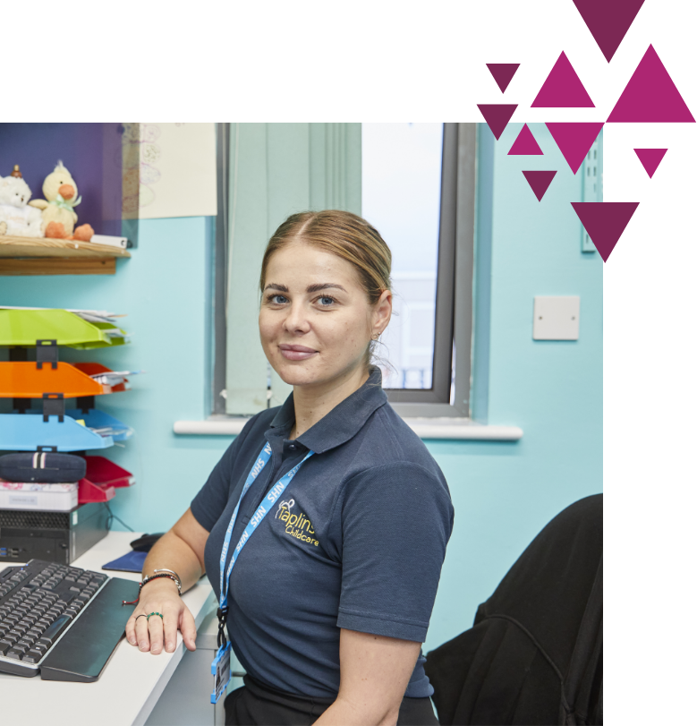 A woman wearing a blue polo shirt and a lanyard sits at a desk with a computer, looking at the camera. the office has a bright window and colorful shelves. pink geometric shapes overlay the top right corner.
