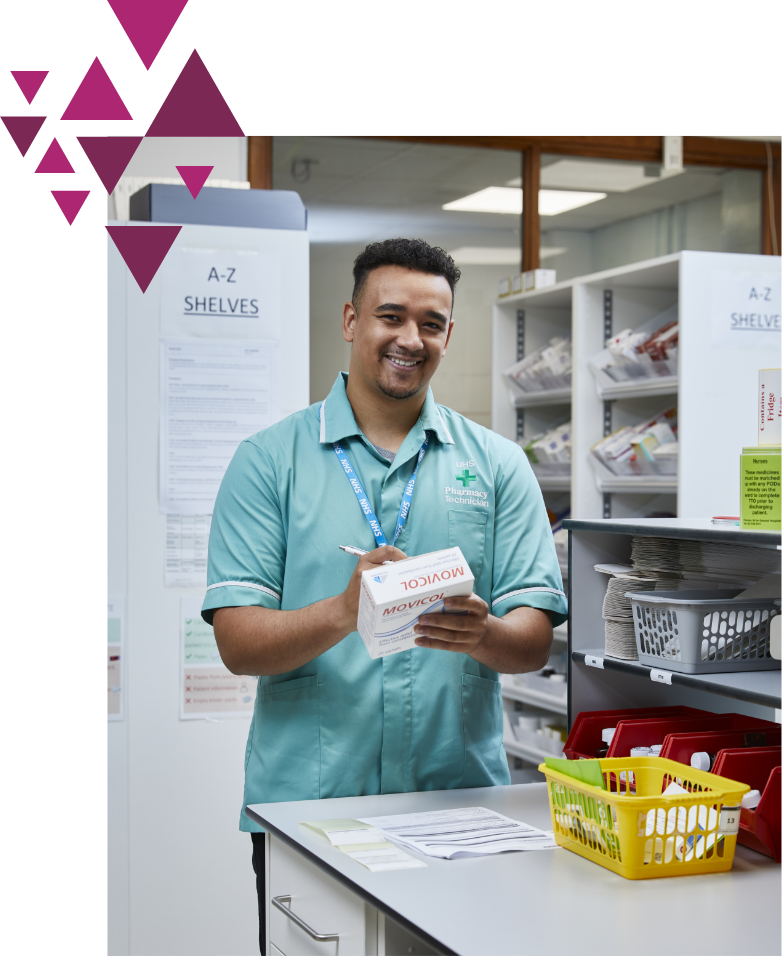 A smiling male pharmacist in a teal uniform stands in a pharmacy, holding a medication box and a prescription form. shelves with various items are visible in the background.