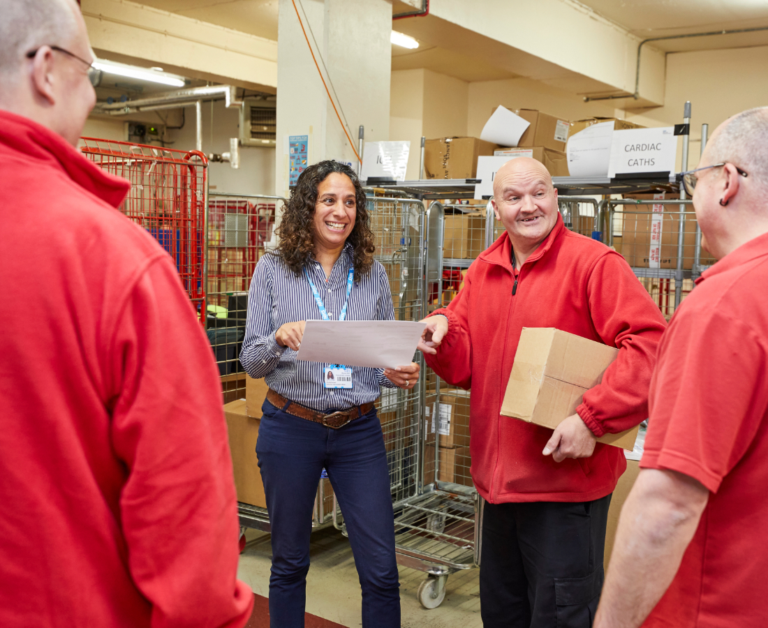 A diverse group of coworkers, one woman and three men, are engaging and smiling in a warehouse. the woman is handing a document to a man who is gesturing enthusiastically.