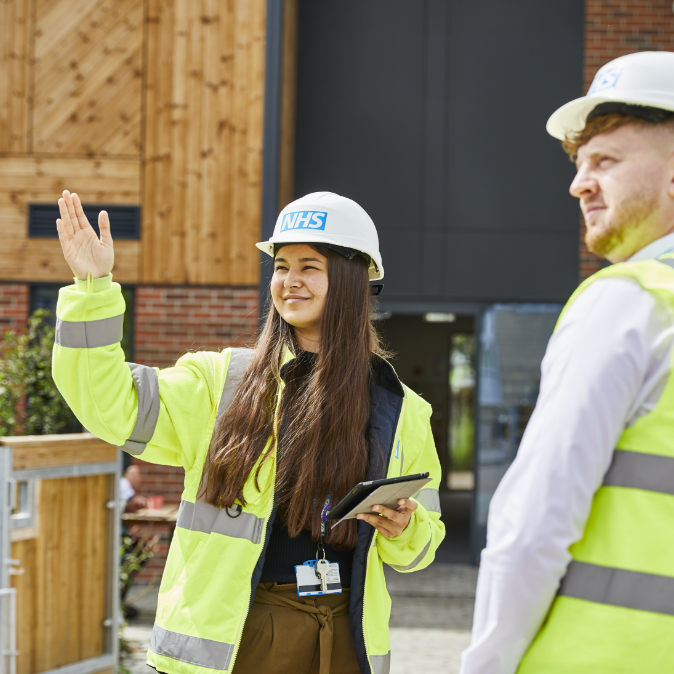 A female construction worker in a high-visibility vest and nhs hard hat gestures while holding a tablet, with a male colleague in the background outside a wooden building.