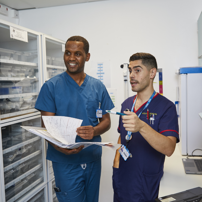Two male healthcare professionals, one holding documents and the other pointing at the papers, are discussing in a medical supply room. both are wearing blue scrubs and id badges.
