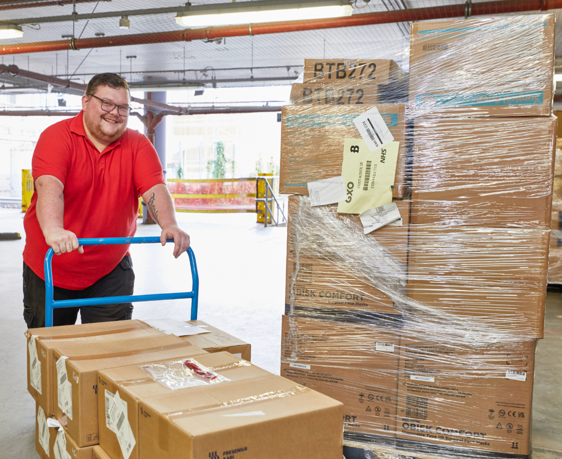 A cheerful worker using a hand truck to transport a stack of cardboard boxes and shrink-wrapped pallets in a warehouse.